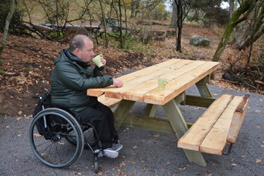 Man i rullstol fikar vid en universellt utformad parkbänk. Bordet har en förlängd bordsskiva så att en rullstol, rollator eller barnvagn kan köra in under.